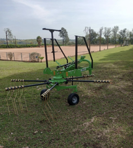 Trator agrícola operando ancinho enleirador em campo de feno, formando leiras uniformes de capim seco; equipamento robusto da Agroforn garante organização e eficiência na fenação.