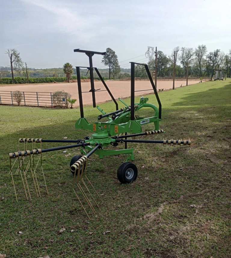 Trator agrícola operando ancinho enleirador em campo de feno, formando leiras uniformes de capim seco; equipamento robusto da Agroforn garante organização e eficiência na fenação.