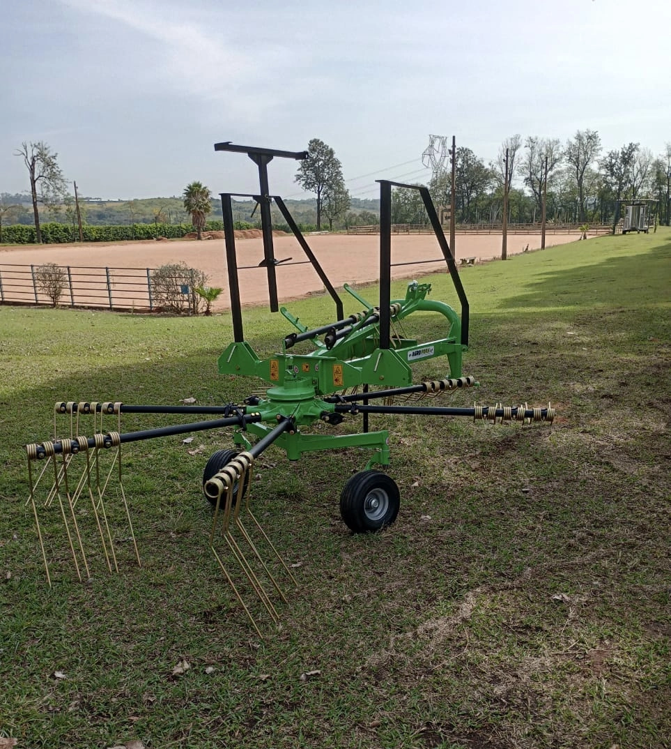 Trator agrícola operando ancinho enleirador em campo de feno, formando leiras uniformes de capim seco; equipamento robusto da Agroforn garante organização e eficiência na fenação.